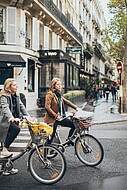 Zwei junge Frauen in Alltagskleigung auf Fahrrädern stehen an einer Straße