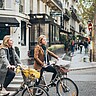 Zwei junge Frauen in Alltagskleigung auf Fahrrädern stehen an einer Straße