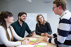 Das Bild zeigt zwei Männer und zwei Frauen, die an einem Tisch stehend miteinander diskutieren.