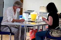 Das Bild zeigt zwei Personen an einem Tisch. Beide tragen eine medizinische Mundnasenbedeckung. Die eine Person hält einen gelben Impfausweis in der Hand und die gegenübersitzende Person füllt einen Zettel aus. Im Hintergrund sind Desinfektionsmittel und andere medizinische Geräte erkennbar. Copyright Pädagogische Hochschule Heidelberg