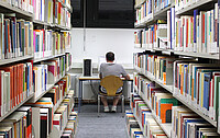 Auf dem Bild sieht man rechts und links Regale mit zahlreichen Büchern. In der Mitte des Bildes sieht man einen jungen Mann von hinten. Er sitzt am Schreibtisch und arbeitet konzentriert. Copyright: PH Heidelberg