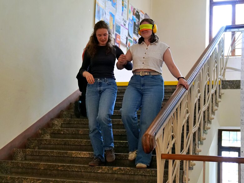Das Foto zeigt zwei Studierende der Taubblindenpädagogik auf der Treppe: Eine führt, die andere trägt eine Augenbinde und Kopfhörer.