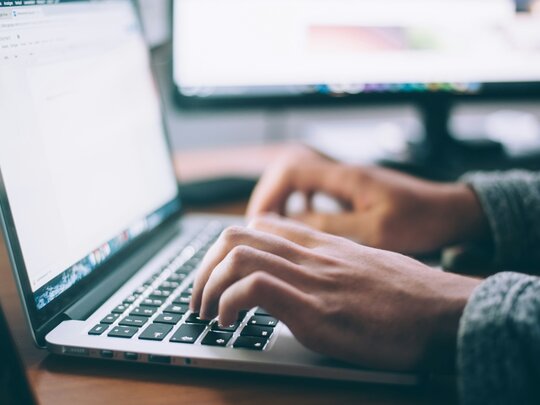 Zwei Hände auf einer Laptoptastatur. Im Hintergrund zwei verschwommene Bildschirme