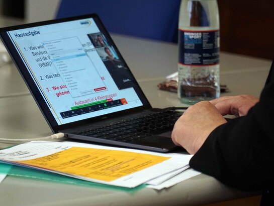 Das Bild zeigt einen Laptop, der auf einem Tisch steht. Eine Person arbeitet an dem Laptop. Um den Laptop liegen Papiere.