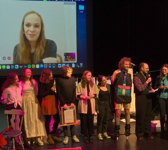 Die studentische Jury steht in einer Reihe und verleiht den Preis der Theatertage. Im Hintergrund ist auf einer Leinwand die Preiträgerin zu sehen