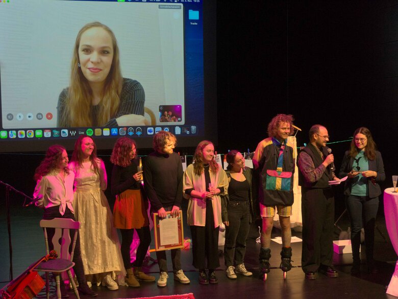 Die studentische Jury steht in einer Reihe und verleiht den Preis der Theatertage. Im Hintergrund ist auf einer Leinwand die Preiträgerin zu sehen