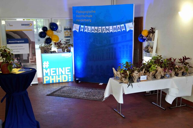 Zu sehen ist die Pflanzenbar. Diese besteht aus einem Alumni-Programm-Rollup, der offiziellen PHHD-Messerückwand ergänzt durch eine Wimpelkette mit dem Schriftzug „Pflanzenbar“, einem Stehtisch, zwei Tischen und einem Leiterregalen die mit ca. 80 kleinen Pflanzen bestückt sind sowie dekorative Lichterketten, blau-gelbe Luftballons und ein Teppich.