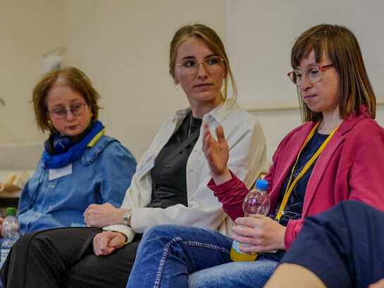Das Bild zeigt Melissa Pfeiffer, die in einem Seminarraum der PH Heidelberg sitzt. Rechts von Frau Pfeiffer sitzt Qualifizierungsteilnehmerin Susann Bensch, links von ihr sitzt Qualifizierungsteilnehmerin Louisa Kabbe. Frau Pfeiffer schaut zu Frau Bensch, die in der Situation spricht.