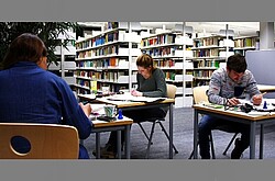 drei Studierende an einzelnen Schreibtischen mit Zetteln, Stiften und Büchern. 