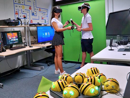 Eine Studentin und ein Student tragen eine VR-Brille auf dem Kopf und Joy-Sticks in den Händen.