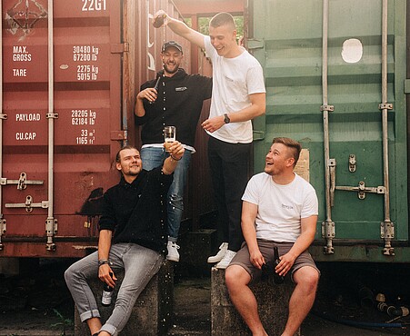 Das Foto zeigt ein Gruppenfoto mit 4 jungen Menschen, die in die Kamera lächeln. Einige von ihnen haben eine Bierflasche in der Hand.