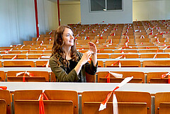 Das Bild zeigt eine Studentin. Sie sitzt alleine in einem Hörsaal und gebärdet mit den Händen. Das Foto wurde an der PH Heidelberg während der Corona-Pandemie aufgenommen; einzelne Sitze sind daher mit Absperrband abgesperrt. Copyright: PH Heidelberg