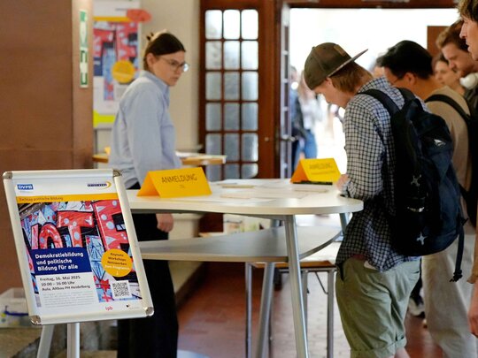 Fokus auf dem Plakat zum Fach Demokratiebildung. Im Hintergrund sieht man unscharf, wie sich Menschen zu dem Tag anmelden.