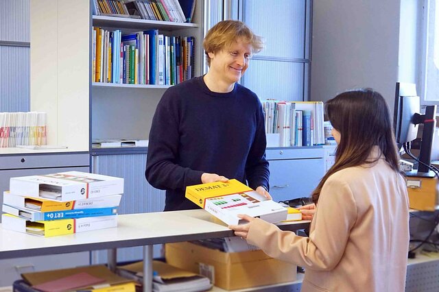 Zu sehen ist ein Mann an einem Tresen, der ein Buch aushändigt.