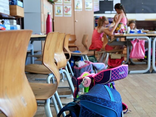 Das Foto zeigt einen Klassenraum. Im Vordergrund sind Schultaschen und Tische und im Hintergrund sind Schüler:innen zu erkennen.
