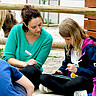 Eine Frau und eine Schülerinnen sitzen im Zoo auf dem Boden. Das Mädchen hält in iPad in der Hand. Die Frau lehnt sich zu ihr rüber.