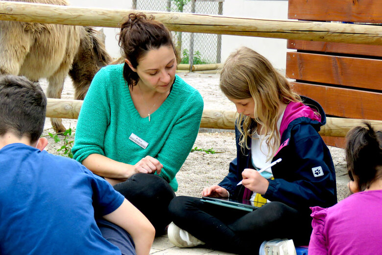 Eine Frau und eine Schülerinnen sitzen im Zoo auf dem Boden. Das Mädchen hält in iPad in der Hand. Die Frau lehnt sich zu ihr rüber.