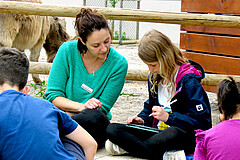 Eine Frau und eine Schülerinnen sitzen im Zoo auf dem Boden. Das Mädchen hält in iPad in der Hand. Die Frau lehnt sich zu ihr rüber.