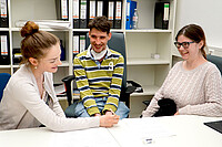 Maria Ritter. Sie sitzt mit Michael Gänßmantel und Anna Neff im Büro und zeigt den beiden etwas auf einem Papier. Alle drei lachen herzlich.