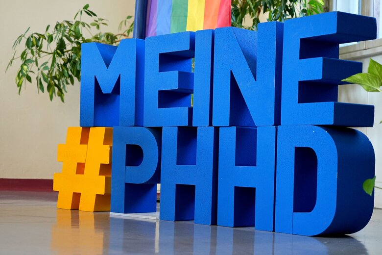 Der Hashtag #MeinePHHD in großen, einzelnen Buchstaben. Dahinter die Diversity-Flagge der Pädagogischen Hochschule Heidelberg.