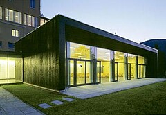 Das Bild zeigt ein Gebäude mit Flachdach und Glasfronten, welches von Grünflächen umgeben ist.
