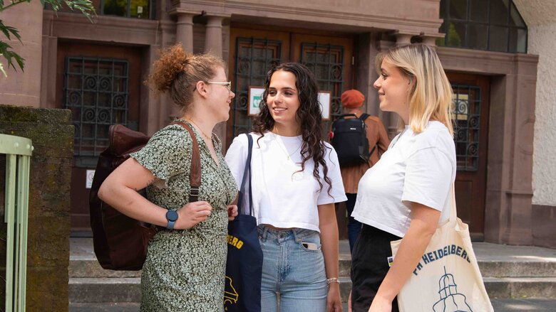 Drei Studentinnen unterhalten sich vor den Haupteingang des Altbaus der Pädagogischen Hochschule Heidelberg.