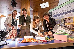 Drei Frauen und ein Mann stehen vor einem Tisch. Auf diesem liegen mehrere Gegenstände aus dem Teilprojekt Climate Change Eduation. Die Personen interagieren mit den Gegenständen. Im Hintergrund sieht man ein wissenschaftliches Poster über das TT-Teilprojekt.