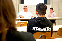 Junger Mann von hinten. Unscharf im Hintergrund sitzen Helmuth Pflantzer und Anna Neff. Das Bild wurde während des Seminars aufgenommen.