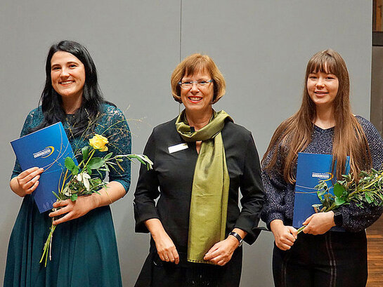Das Bild zeigt die Preisträgerinnen Gina-Maria Trunte und Janine Kreschel. Beide halten eine blaue Mappe und eine gelbe Rose in der Hand. Zwischen ihnen steht Dr. Monika Mölders-Felgenhauer
