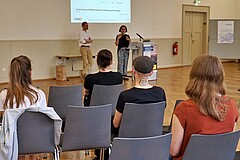Das Bild zeigt zwei Redner:innen, die an einem Pult einen Vortrag halten. Ihre Zuschauer:innen sind von hinten abgebildet.