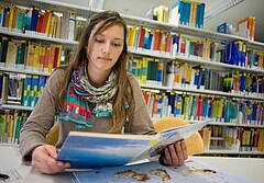 Das Bild zeigt im Vordergrund eine Studentin, welche in der Bibliothek sitzt und liest. Im Hintergrund ist ein mit zahlreichen Büchern bestücktes Regal zu sehen.