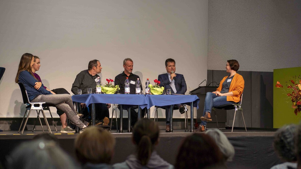 Sechs Menschen sitzen im Rahmen der Podiumsdiskussion auf der Bühne der Mehrzweckhalle. 