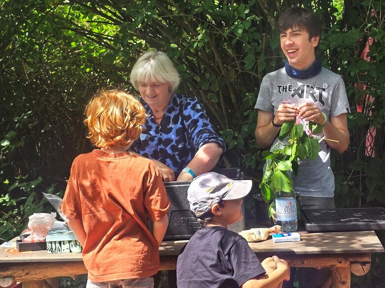 Lissy Jäkel arbeitet mit einem jungen Mann im Ökogarten an einem Tisch. Zwei Kinder sind mit dem Rücken zu sehen.
