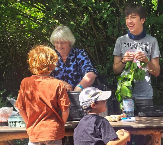Lissy Jäkel arbeitet mit einem jungen Mann im Ökogarten an einem Tisch. Zwei Kinder sind mit dem Rücken zu sehen.