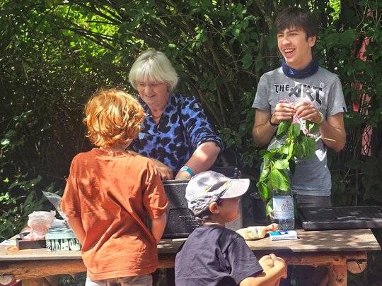 Lissy Jäkel arbeitet mit einem jungen Mann im Ökogarten an einem Tisch. Zwei Kinder sind mit dem Rücken zu sehen.