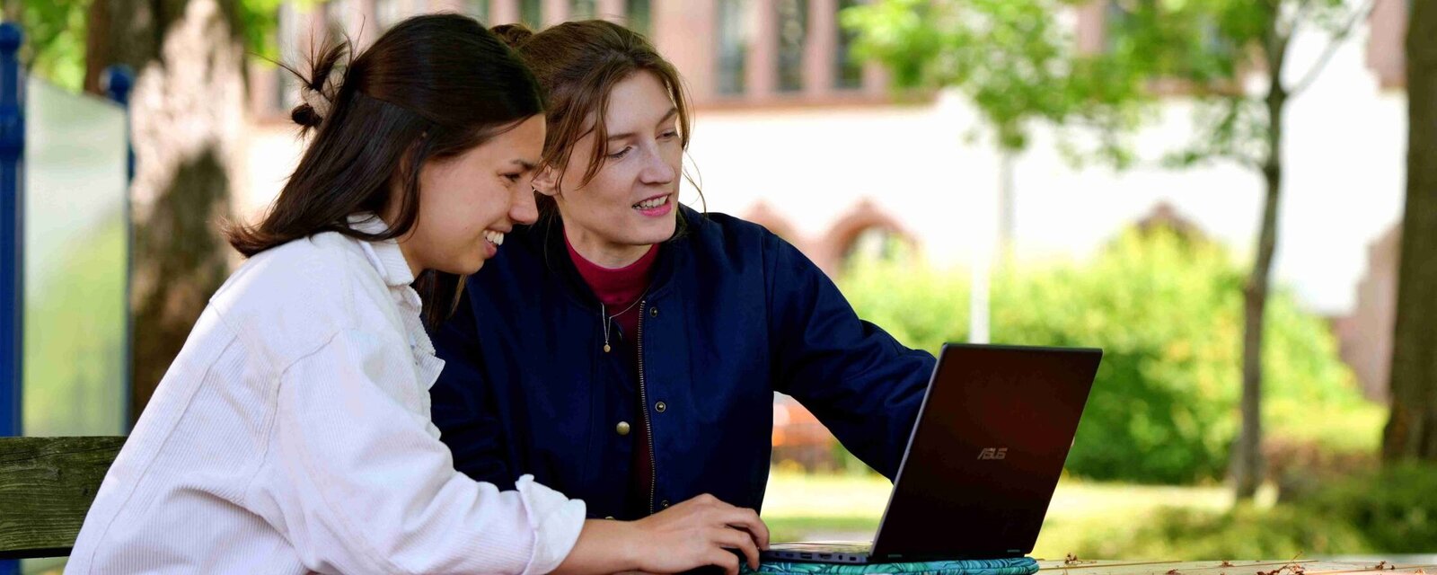 Zwei Studentinnen sitzen auf dem Campus im Innenhof des Altbaus am Laptop und informieren sich über digitale Studien- und Prüfungsorganisation im CAMPUS-Portal.