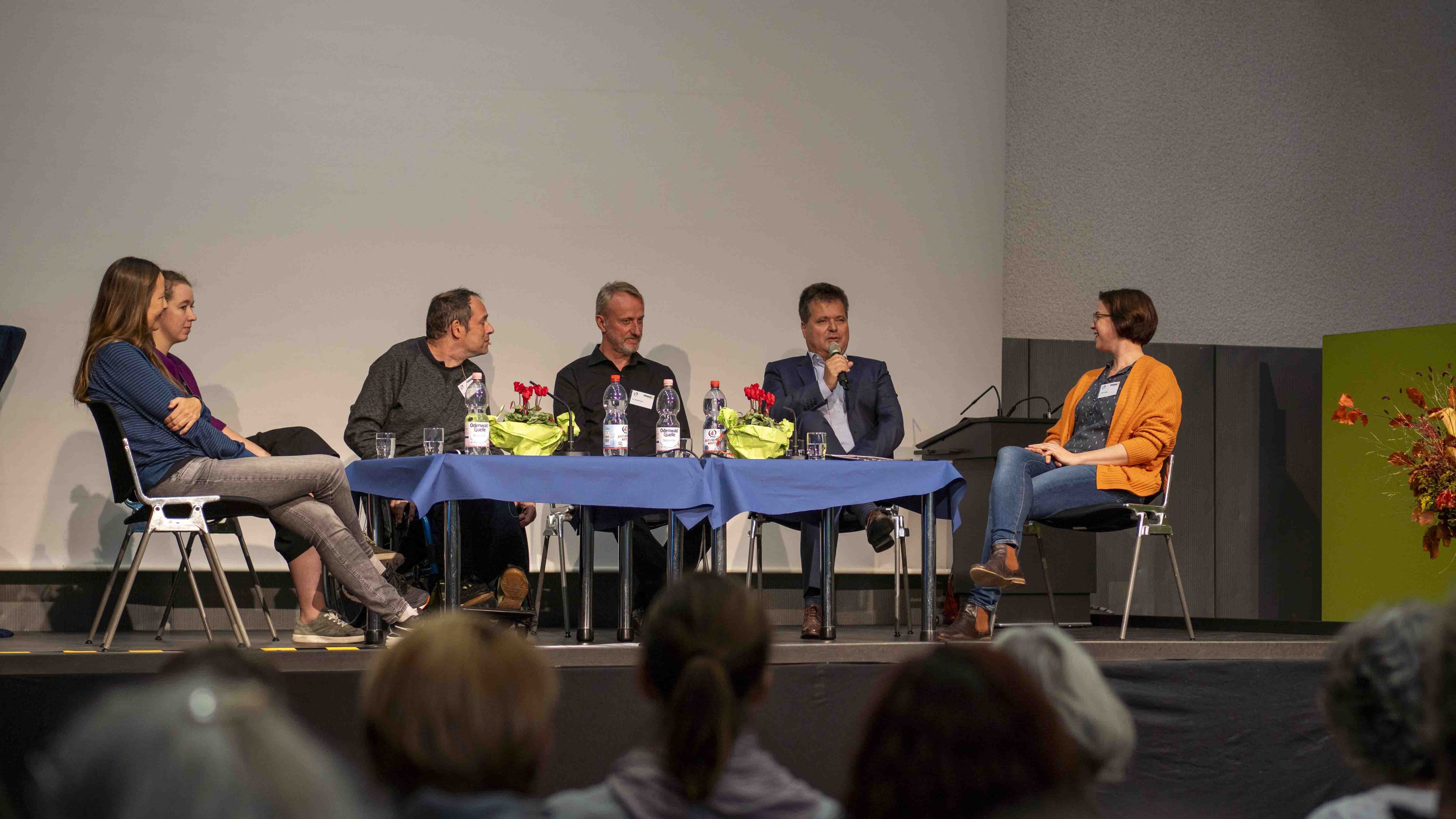 Sechs Menschen sitzen im Rahmen der Podiumsdiskussion auf der Bühne der Mehrzweckhalle. 
