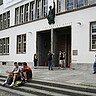 Das Bild zeigt die Neue Universität von außen. Davor sitzen und stehen eine Vielzahl von Studierenden.
