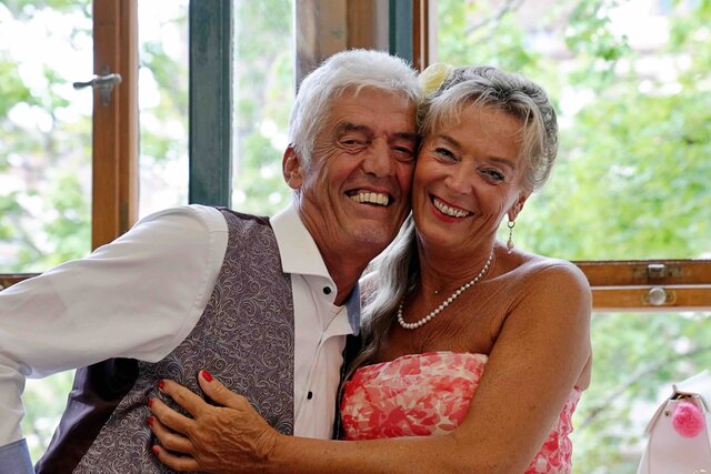 Zu sehen sind zwei Gäste (Frau rechts, Mann links) beim Sektempfang, die sich umarmen und lachend in die Kamera schauen.