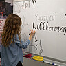 Eine junge Frau schreibt "herzlich willkommen" an ein Whiteboard. Links danebn steht ein Roll-Up von der ZOrA-Akademie.