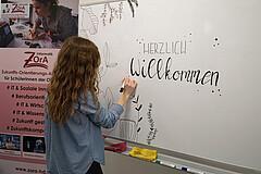 Eine junge Frau schreibt "herzlich willkommen" an ein Whiteboard. Links danebn steht ein Roll-Up von der ZOrA-Akademie.