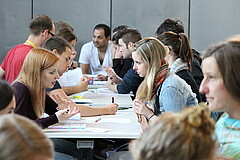 Das Bild zeigt sich an Tischen gegenübersitzende Studierende, die Unterhaltungen führen.rhalten.