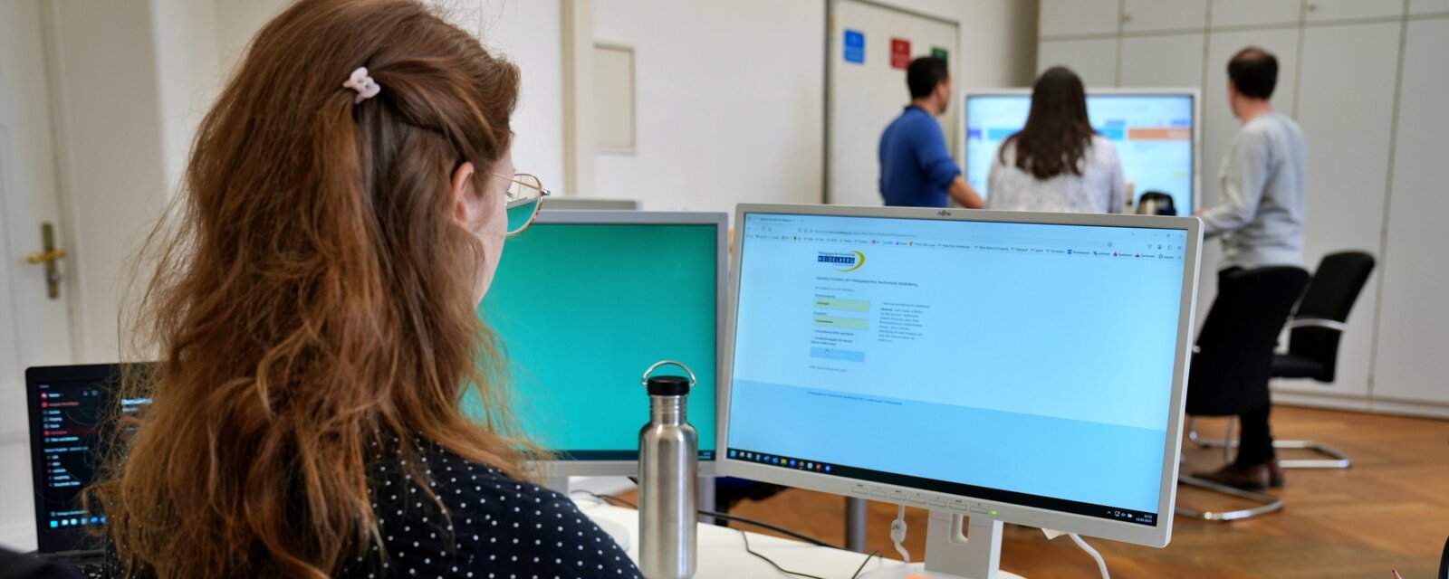 Mitarbeitende im Einsatz beim Arbeiten an der PHHD: Im Vordergrund ist eine Frau zu sehen, die vor dem PC sitzt. Im Hintergrund stehen drei Personen an einem Steharbeitstisch und blicken auf einen Bildschirm. Sie sind unscharf zu sehen.
