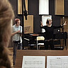 Das Bild zeigt Heike Kiefner-Jesatko im Studio. Sie dirigiert den Chor mit den Händen. Neben ihr steht ein junger Mann. Er trägt Kopfhörer und schaut auf einen Laptop. Vorne links sieht man eine der Sängerinnen von schräg hinten. Es handelt sich um einen Screenshot aus dem Promovideo für das Album.