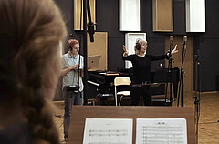 Das Bild zeigt Heike Kiefner-Jesatko im Studio. Sie dirigiert den Chor mit den Händen. Neben ihr steht ein junger Mann. Er trägt Kopfhörer und schaut auf einen Laptop. Vorne links sieht man eine der Sängerinnen von schräg hinten. Es handelt sich um einen Screenshot aus dem Promovideo für das Album.