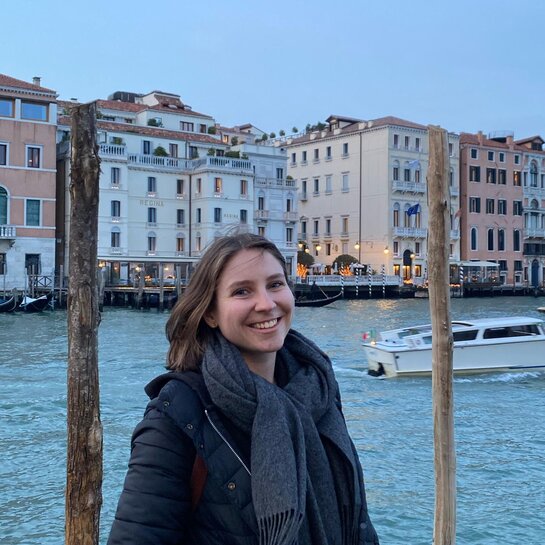 Charlotte steht in Winterklamotten vor einem Fluss, auf dem ein Schiff fährt. Auf der anderen Uferseite ist eine Häuserreihe zu erkennen und eine beleuchtete Straße.