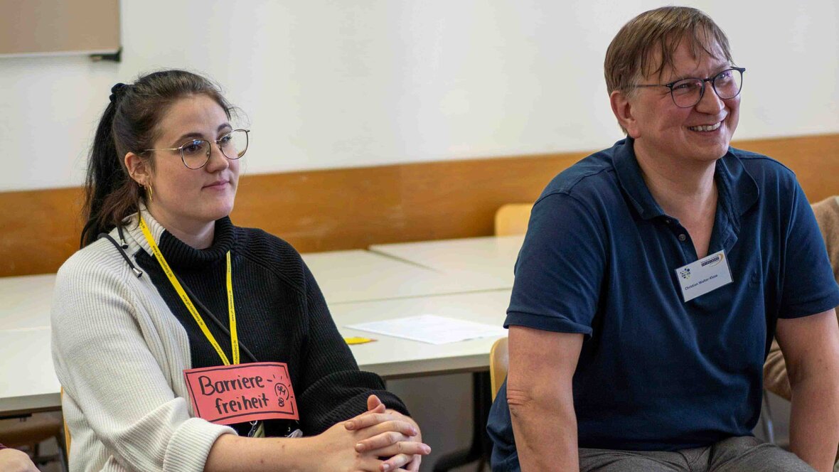 Zwei Personen hören einem Beitrag zu. Eine Frau hat ein Schild um. Darauf steht: "Barrierefreiheit"