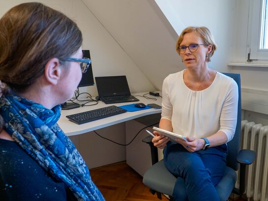 Das Bild zeigt Stephanie Schleer, die in ihrem Büro mit Bildungsfachkraft Anna Neff spricht. Frau Schleer hält ein Tablet in der Hand.