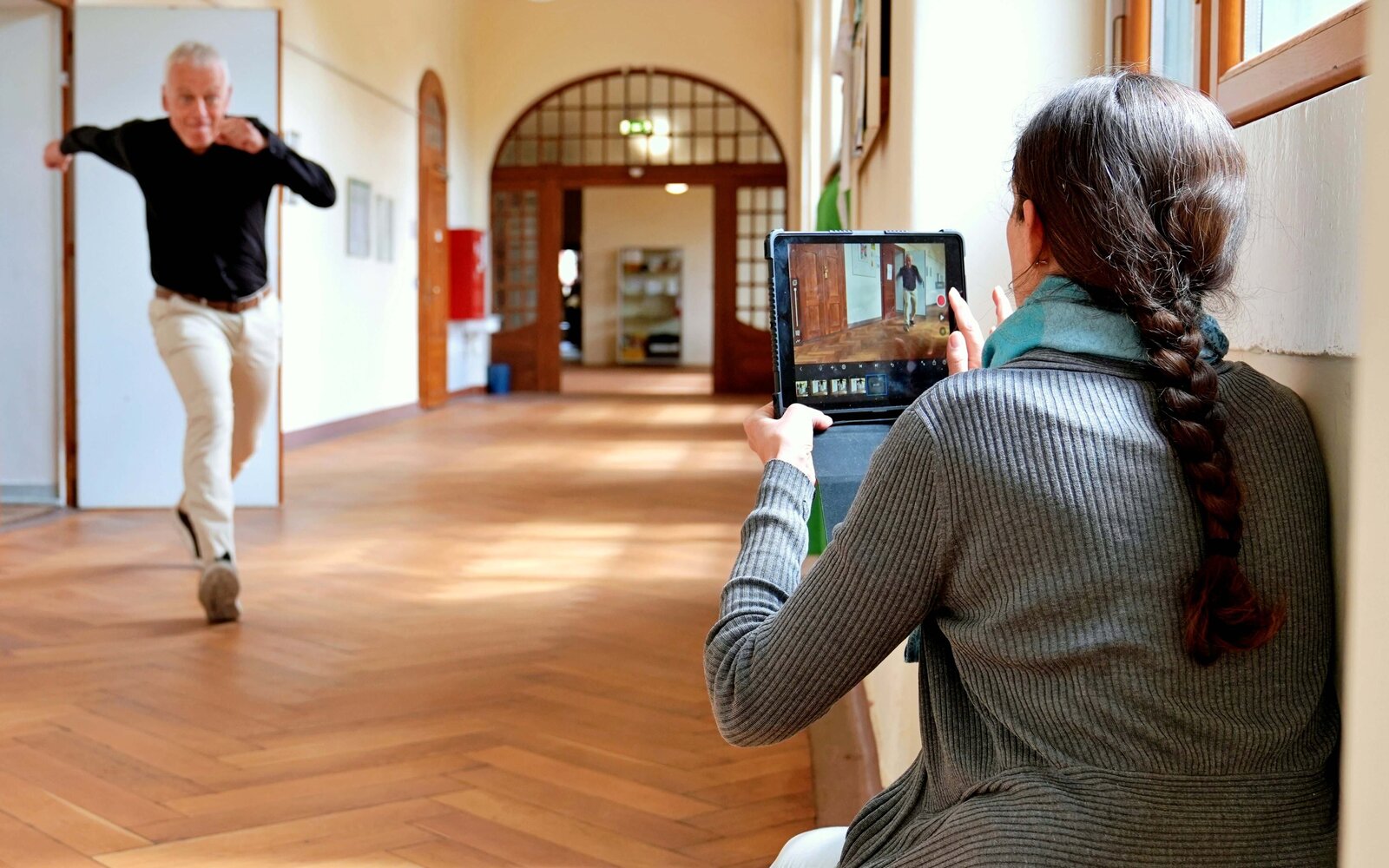 Aufnahme aus dem Altbauflur der PHHD. Ein Frau hockt auf dem Boden und hält ein Tablet hoch. Mit diesem filmt sie einen Mann. Dieser ist unscharf zu sehen, wie er schwungvoll aus einem Raum läuft.