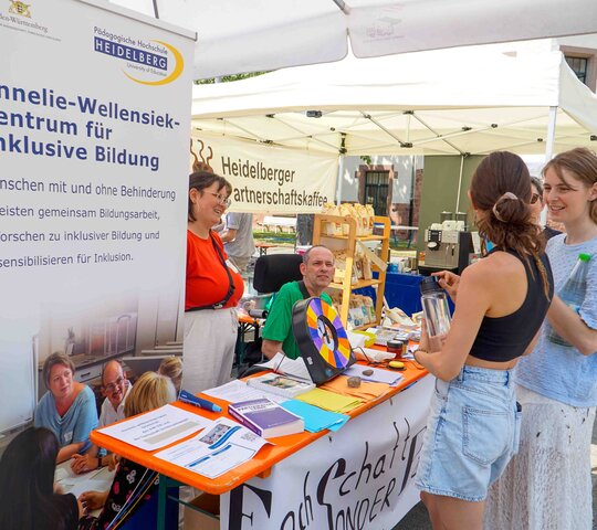 Das Foto zeigt den Stand des AW-ZIB und der Fachschaft Sonderpädagogik bei den PH Days. Im Vordergrund ist ein langer Tisch, auf dem sich verschiedeneDinge befinden: das Glücksrad, bunte Karten, auf denen die Fragen geschrieben stehen, Preise. Vor dem Tisch stehen zwei Studentinnen, hinter dem Tisch sind zwei Personen, davon sitzt eine Person im Rollstuhl. Links neben dem Stand ist ein großer Banner. Auf disem steht: Annelie-Wellensiek-Zentrum für Inklusive Bildung.